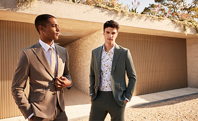 Blog Tile Preview - Two men wearing POLITIX Spring Summer Collection tailored suits – sage green suit with floral shirt and mushroom tailored suit – styled outdoors in modern architecture setting.